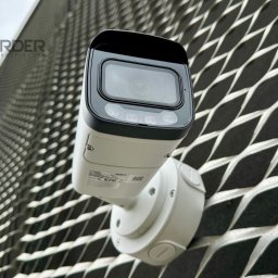 Rekorder.pl - Monitoring i Systemy Smart - Kamera monitoringu IP, biała, zamontowana na metalowej elewacji o strukturze rombów. Widok z dołu, logo firmy w lewym górnym rogu. Obiektyw skierowany w górę.