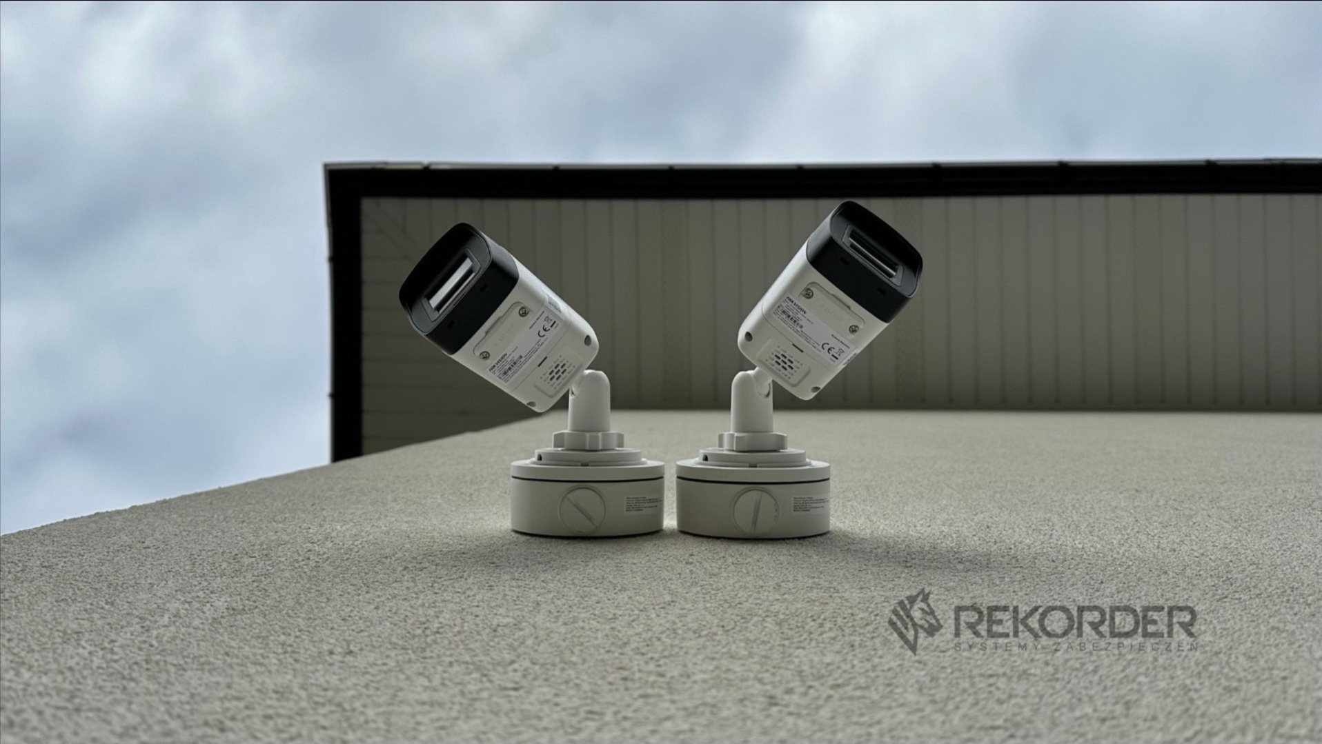 Dwie kamery monitoringu Hikvision zamontowane na elewacji budynku, skierowane w różne strony. Widoczne logo firmy Rekorder na dole zdjęcia.