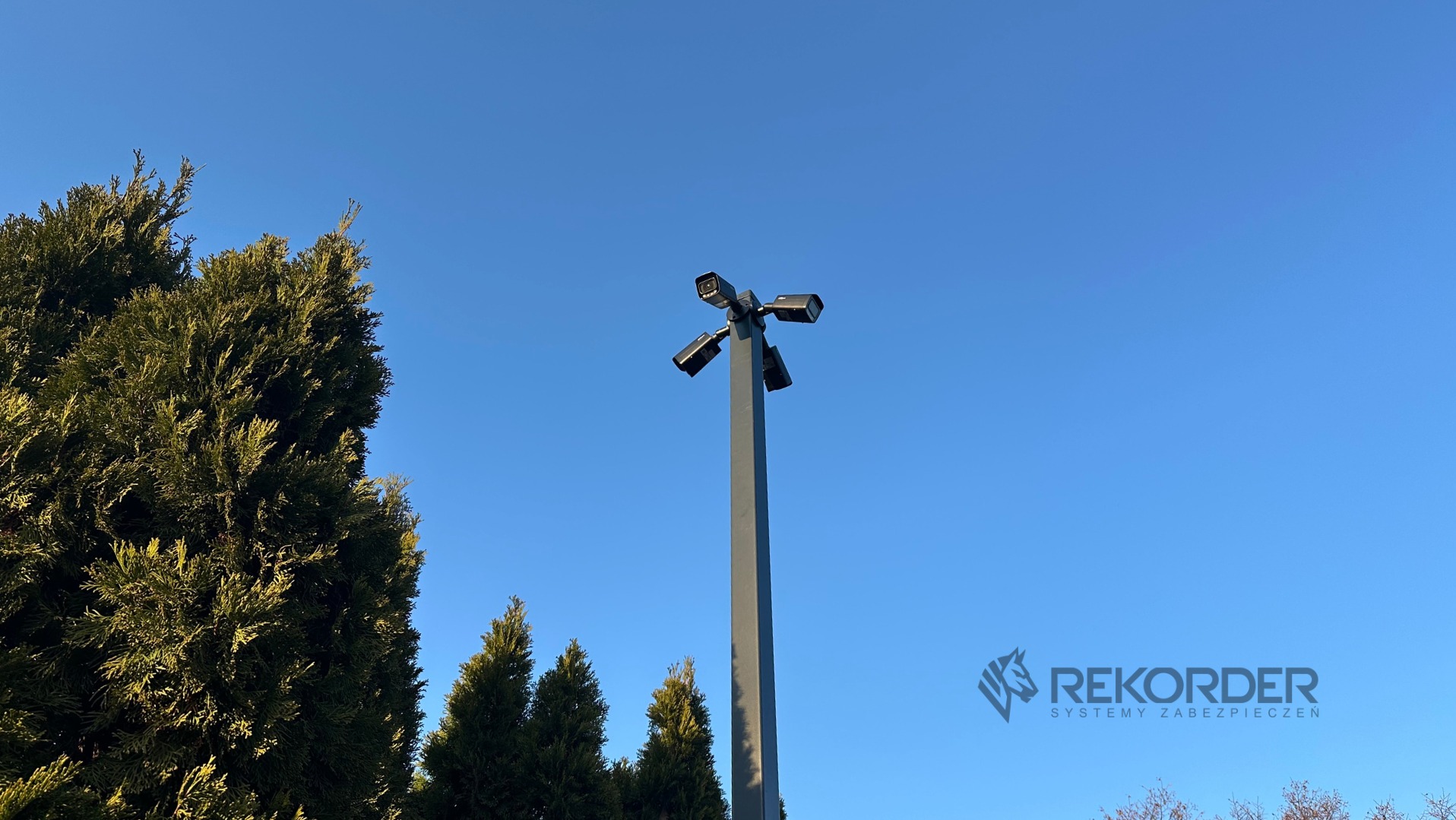 Cztery kamery monitoringu na wysokim słupie, skierowane w różne strony, na tle błękitnego nieba i zielonych drzew. Widoczne logo firmy Rekorder Systemy Zabezpieczeń.