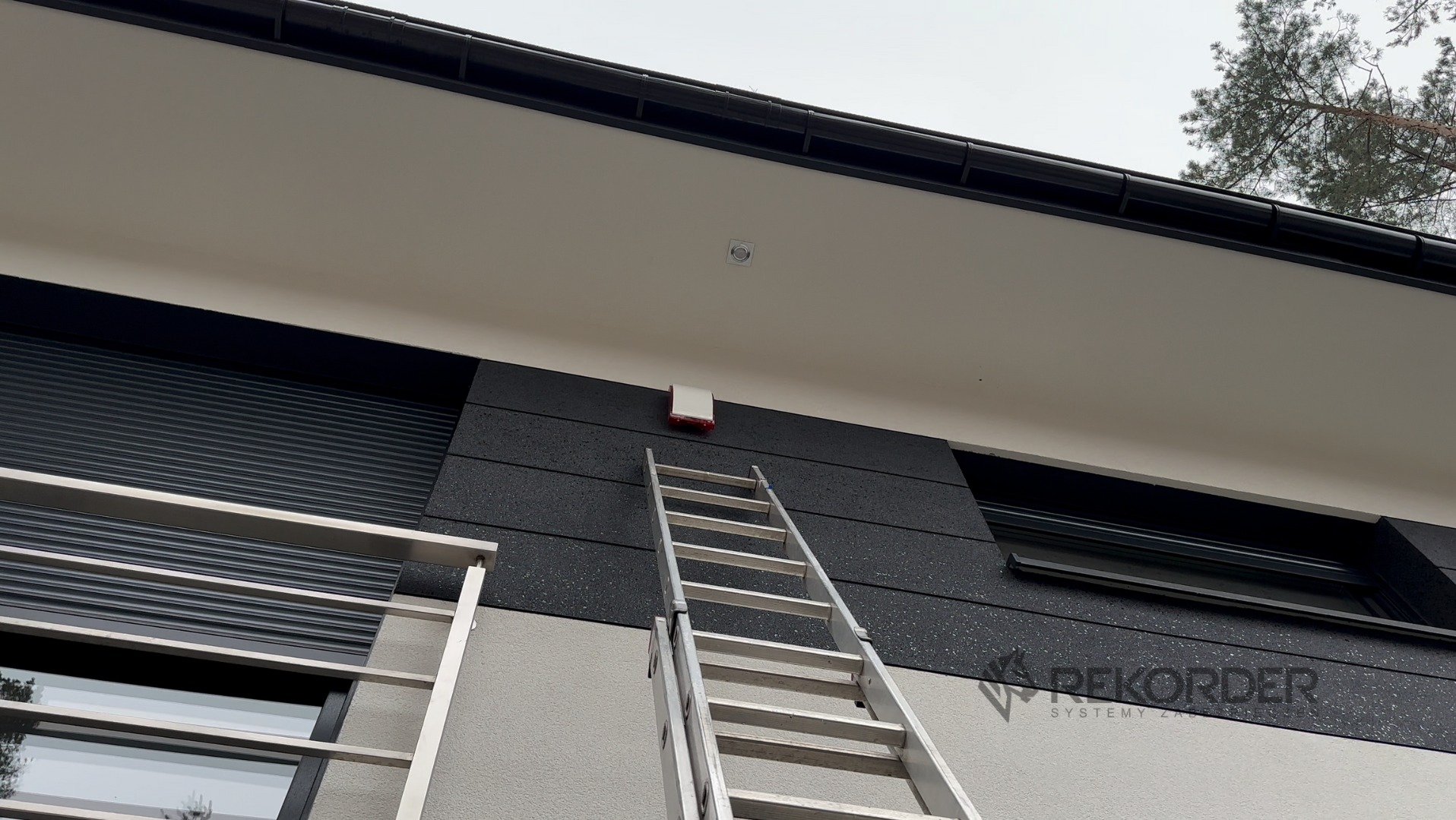 Instalacja czujnika alarmowego na elewacji domu z użyciem drabiny. Widoczny fragment okna z roletą zewnętrzną i logo firmy na ścianie.