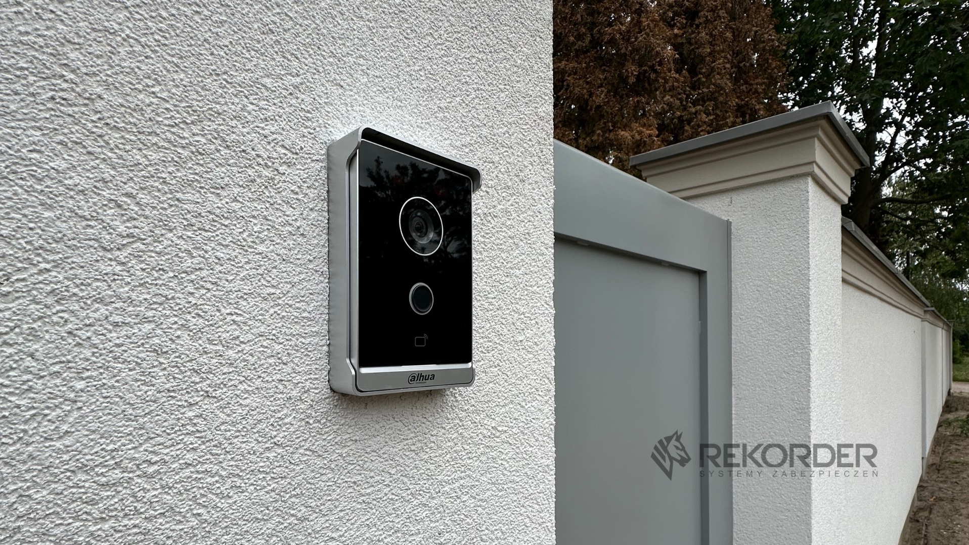 Nowoczesny wideodomofon Dahua zamontowany na teksturowanej, białej ścianie obok szarego ogrodzenia z dekoracyjnymi elementami. Widoczne logo firmy Rekorder.