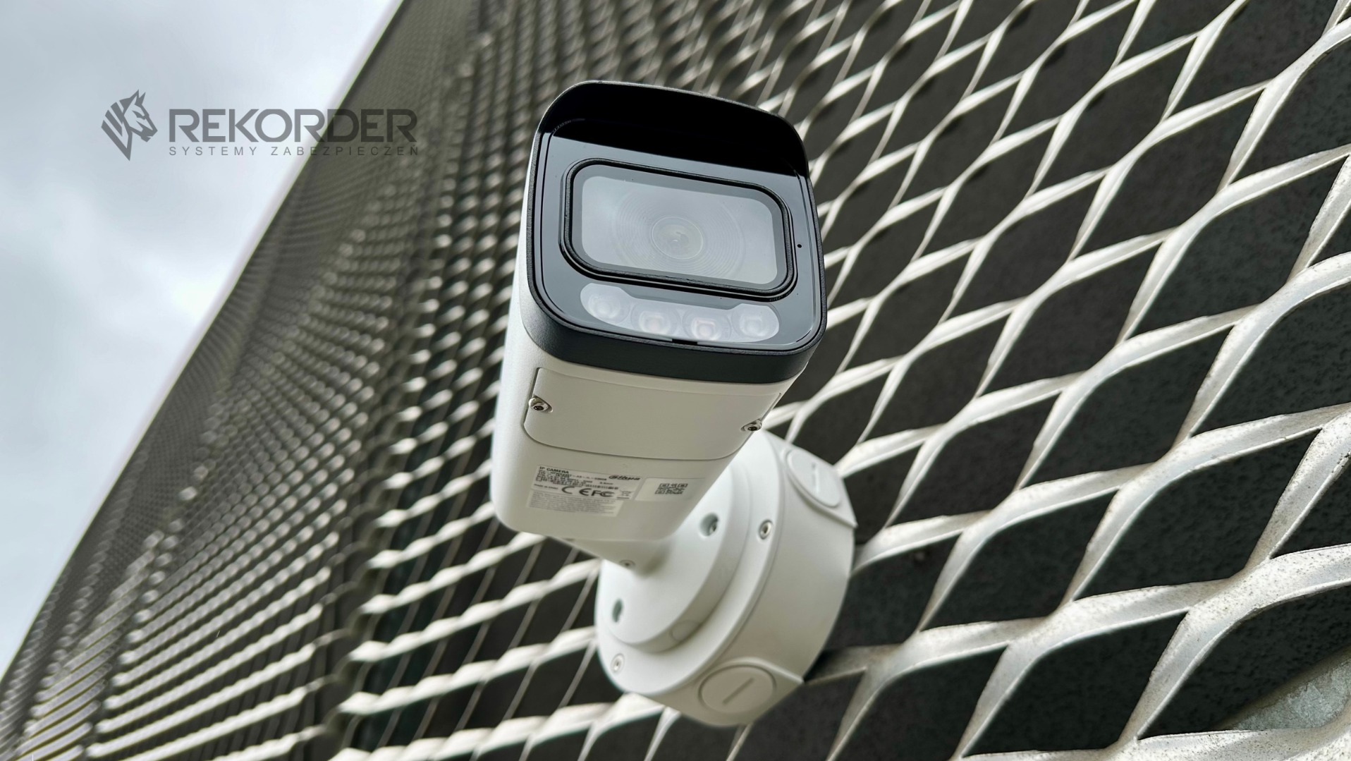 Kamera monitoringu IP, biała, zamontowana na metalowej elewacji o strukturze rombów. Widok z dołu, logo firmy w lewym górnym rogu. Obiektyw skierowany w górę.