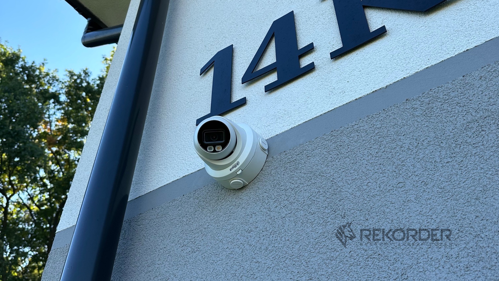 Kamera IP Dahua zamontowana na elewacji budynku obok numeru 141, z widoczną rynną. Ujęcie z dołu, w tle błękitne niebo i zieleń drzew.
