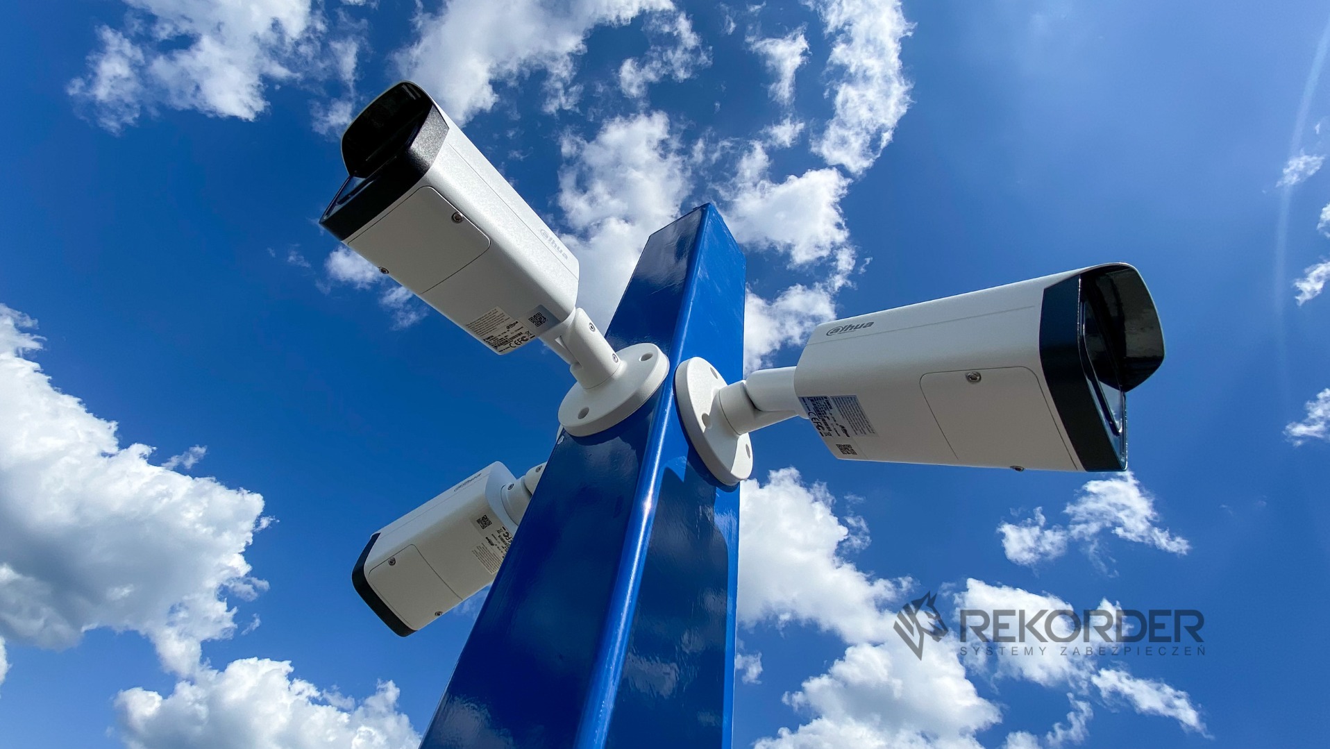 Cztery kamery monitoringu Dahua na niebieskim słupie, skierowane w różne strony, na tle błękitnego nieba z chmurami. Widok z dołu, logo firmy w prawym dolnym rogu.