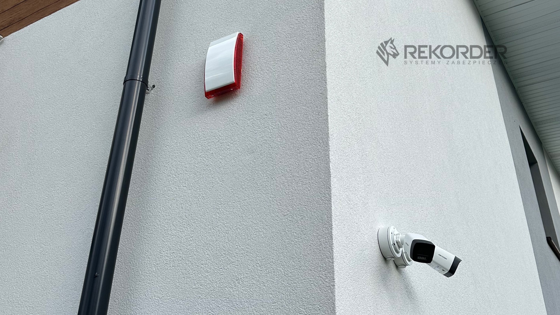 Zewnętrzny montaż: kamera IP Hikvision, sygnalizator alarmowy i rura spustowa na elewacji budynku. Widoczne logo firmy Rekorder Systemy Zabezpieczeń.