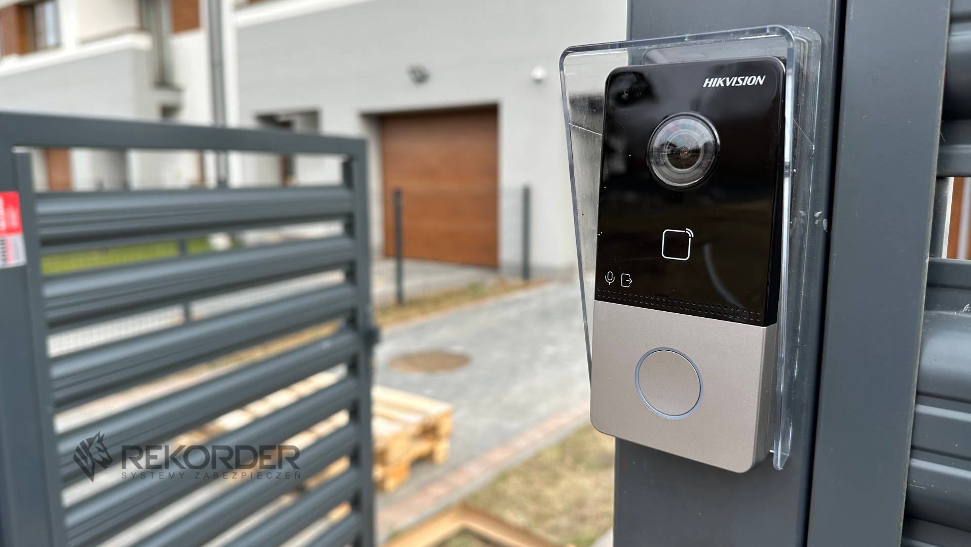 Zewnętrzny panel wideodomofonu Hikvision zamontowany na metalowej furtce, z widocznym budynkiem w tle. Ochronna osłona z tworzywa.