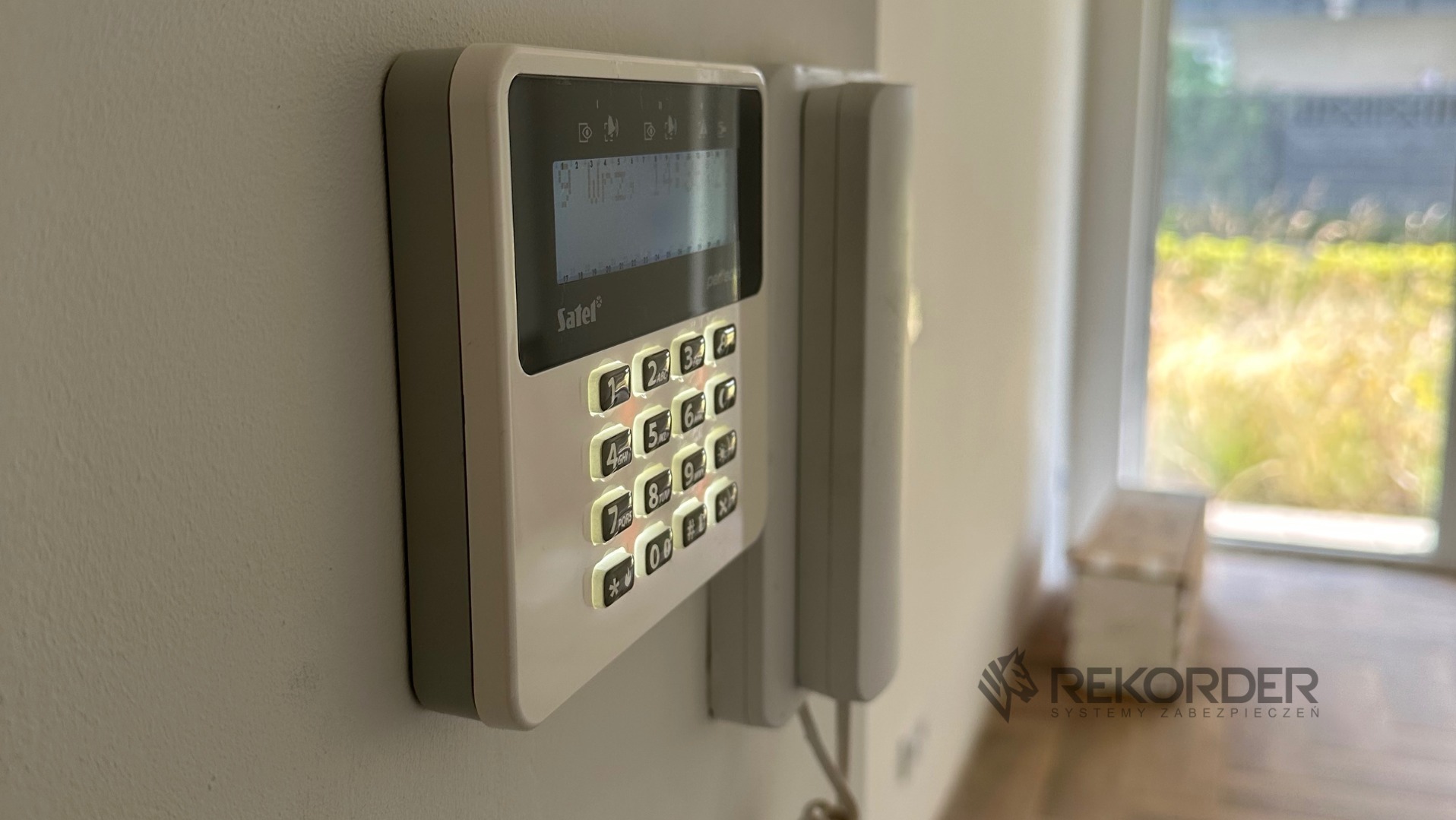 Panel sterowania alarmem Satel Perfecta z podświetlanymi przyciskami, zamontowany na ścianie w jasnym wnętrzu, obok domofon. Widoczne logo firmy na dole.
