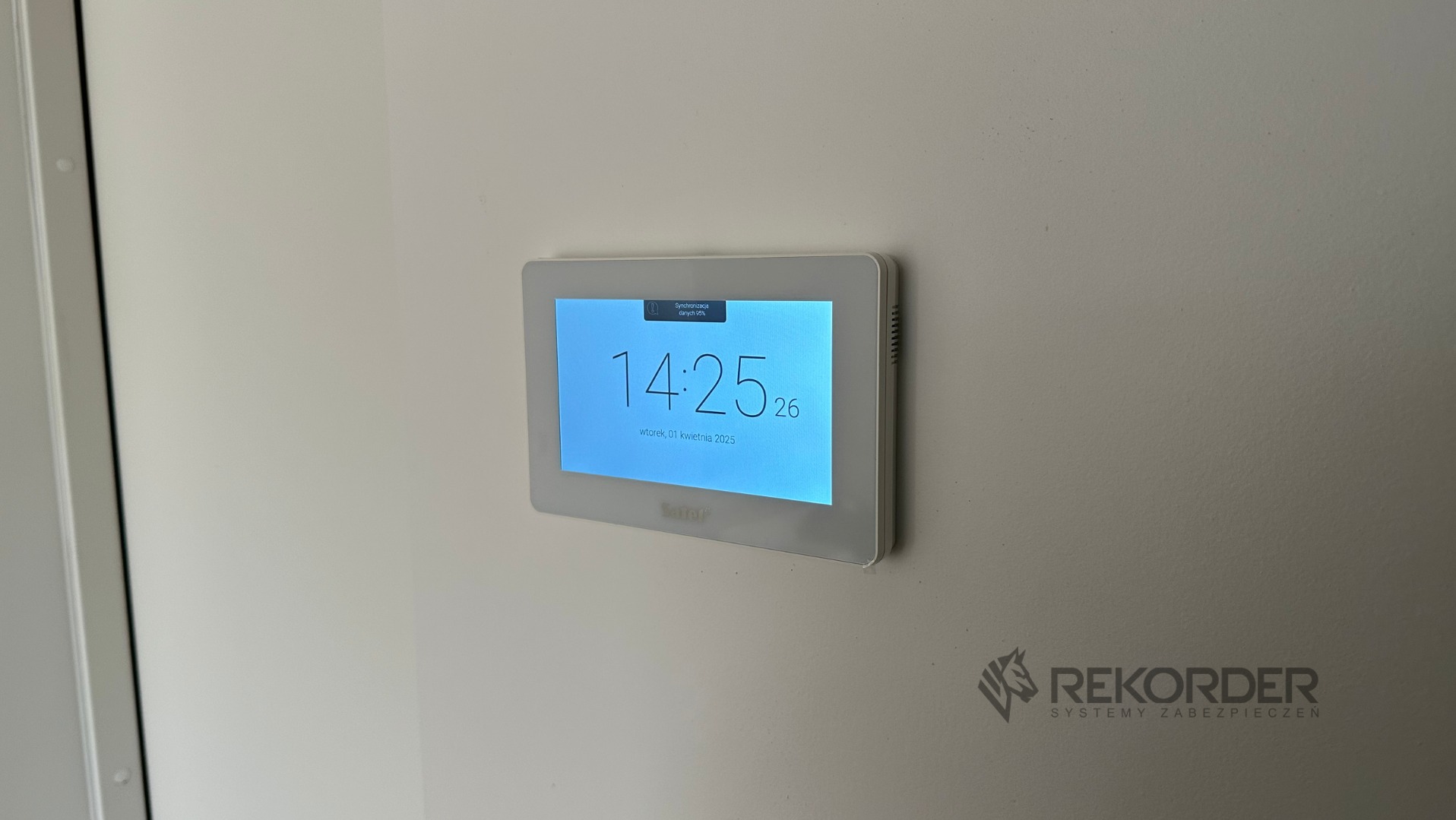 Panel sterowania systemu alarmowego na ścianie, wyświetlający godzinę 14:25 i datę 1 kwietnia 2025. Widoczne logo firmy Rekorder Systemy Zabezpieczeń.