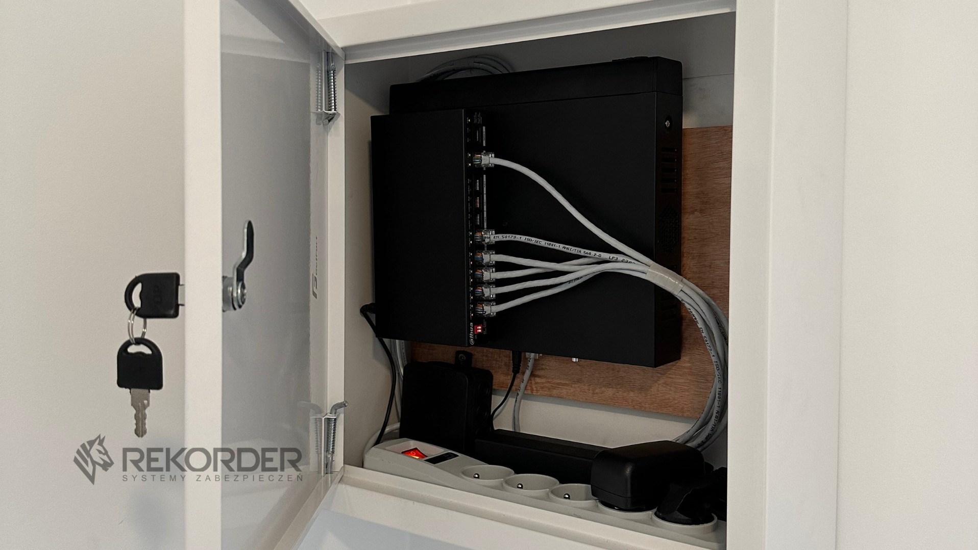 Szafka z centralą alarmową, kablem Ethernet i listwą zasilającą. Klucze do szafki wiszą na drzwiczkach. Widoczne logo firmy Rekorder Systemy Zabezpieczeń.