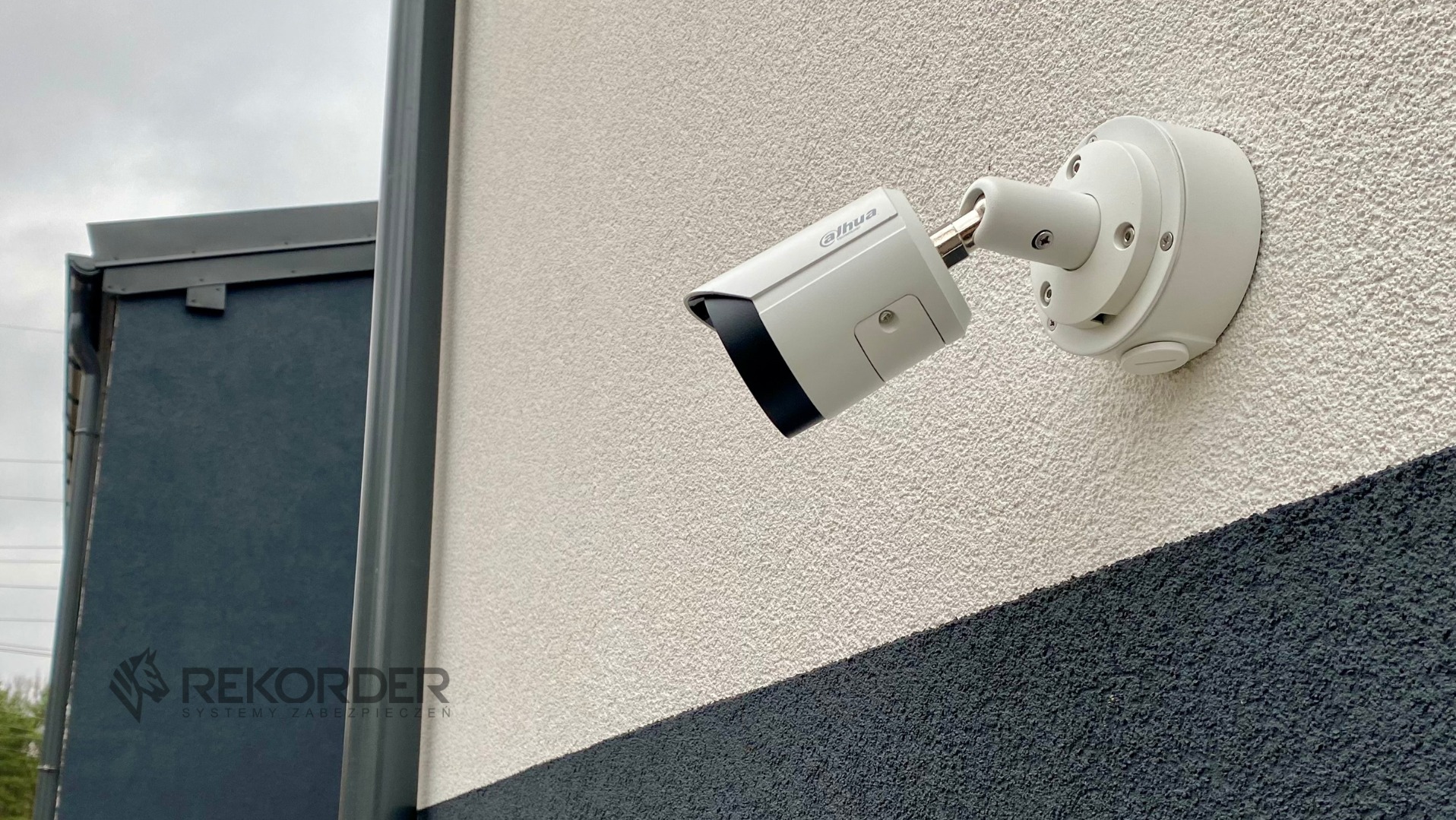 Kamera monitoringu Dahua zamontowana na elewacji budynku z widoczną fakturą tynku i fragmentem ciemnej ściany obok. Widoczne śruby mocujące.
