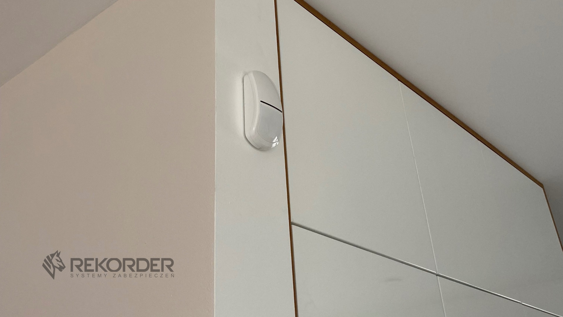 Biały czujnik ruchu zamontowany na ścianie obok białej szafy z drewnianymi akcentami. Widoczne logo firmy Rekorder Systemy Zabezpieczeń.