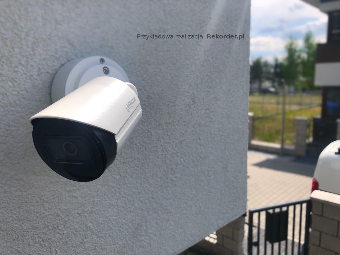 Biała kamera monitoringu Dahua zamontowana na szarej, teksturowanej ścianie zewnętrznej budynku, widok z boku, z fragmentem ogrodzenia i zieleni w tle.