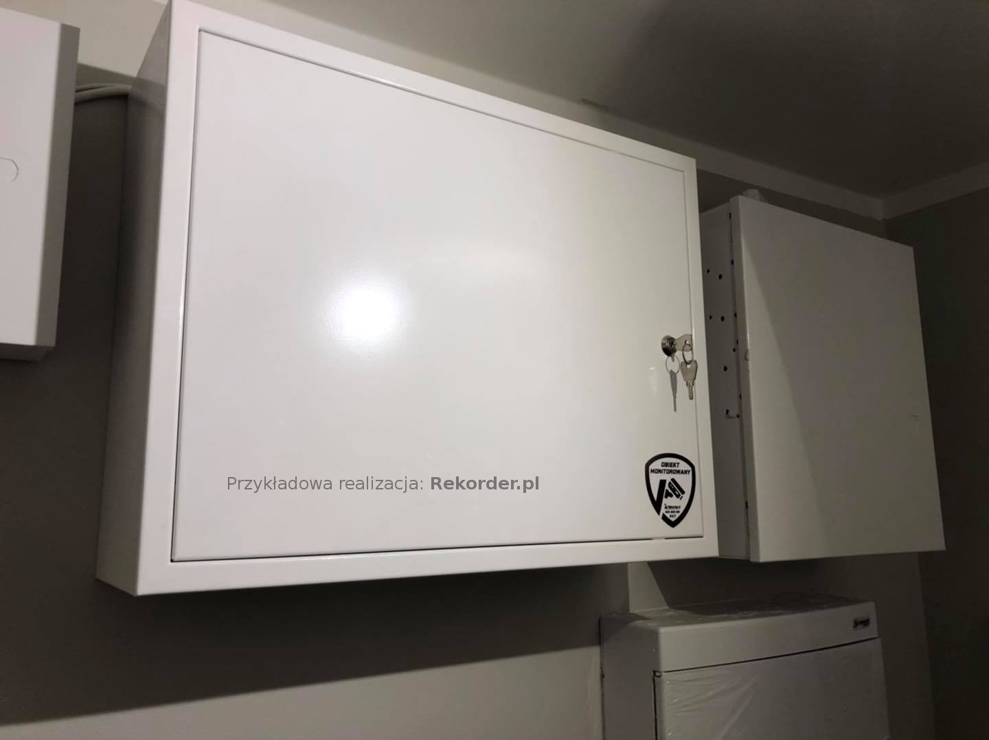 Biała skrzynka na urządzenia monitoringu, zamontowana na ścianie, z widocznymi kluczami i naklejką 'Obiekt Monitorowany', podpisana 'Przykładowa realizacja: Rekorder.pl'.