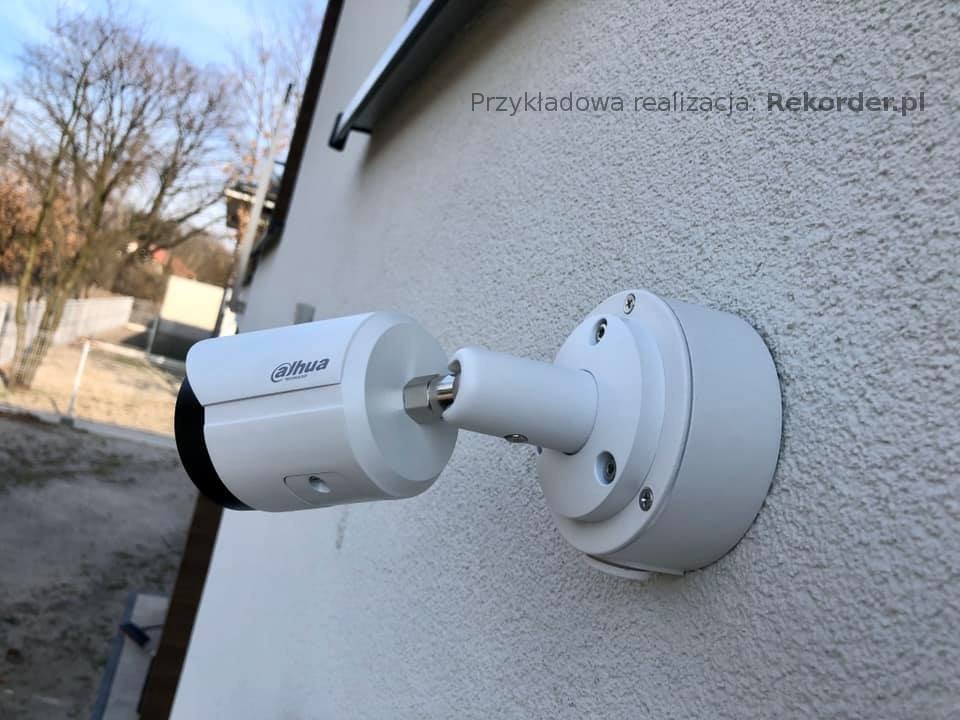 Biała kamera Dahua zamontowana na szorstkiej, jasnej ścianie budynku, widoczne śruby mocujące i fragment dachu oraz drzewa w tle.