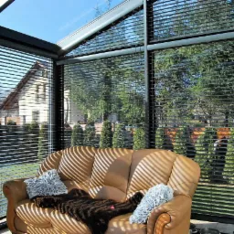 Wnętrze przeszklonego pomieszczenia z aluminiowymi żaluzjami w kolorze antracytu, przez kt&oacute;re widać dom i zieleń. Na pierwszym planie sk&oacute;rzana sofa z poduszkami i narzutą, oświetlona promieniami...