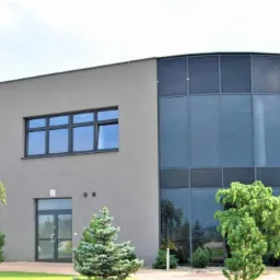 Nowoczesny budynek biurowy z szarymi ścianami i dużymi, ciemnymi oknami, w tym panoramiczne okno narożne, otoczony zielenią.