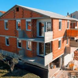 Omega Top Sp. z o.o. - Surowy stan budynku wielorodzinnego z częściowo zamontowanymi oknami i drzwiami balkonowymi, widoczne cegły i beton, robotnicy na dachu.