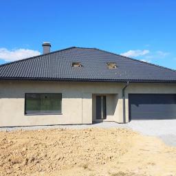 Omega Top Sp. z o.o. - Nowoczesny dom jednorodzinny z grafitową dachówką i ciemnoszarą stolarką okienną, widok z przodu na tle błękitnego nieba. Okno w salonie odbija krajobraz.