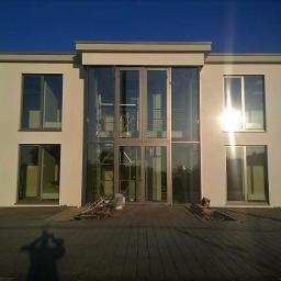 Omega Top Sp. z o.o. - Nowoczesny budynek z dużymi oknami i szklanym wejściem, widoczne rusztowanie wewnątrz, cień osoby w dolnej części kadru.