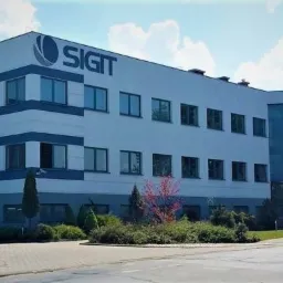 Nowoczesny budynek biurowy z licznymi oknami z ciemnymi ramami, logo firmy na fasadzie, otoczony zielenią i chodnikiem.