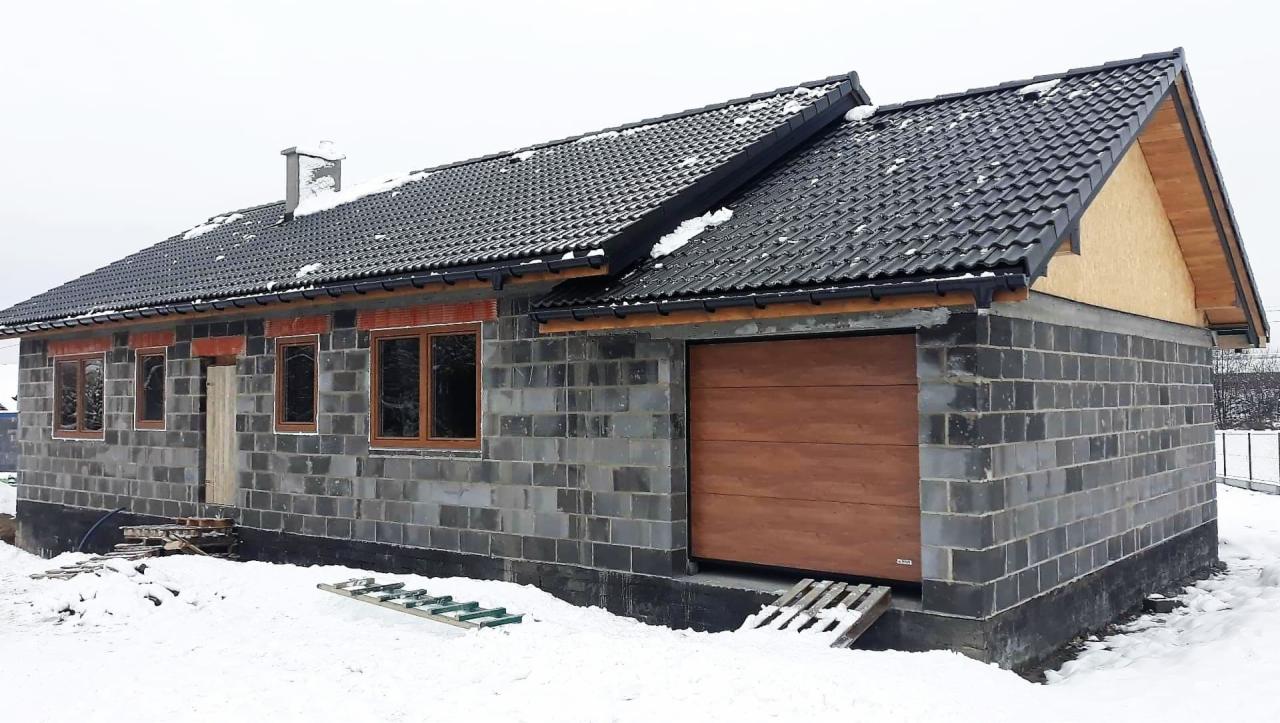 Budynek w stanie surowym zamkniętym, z zamontowanymi brązowymi oknami i drewnianą bramą garażową, ośnieżony dach z czarnej dachówki, ściany z szarego bloczka betonowego, widoczny fragment drabiny.