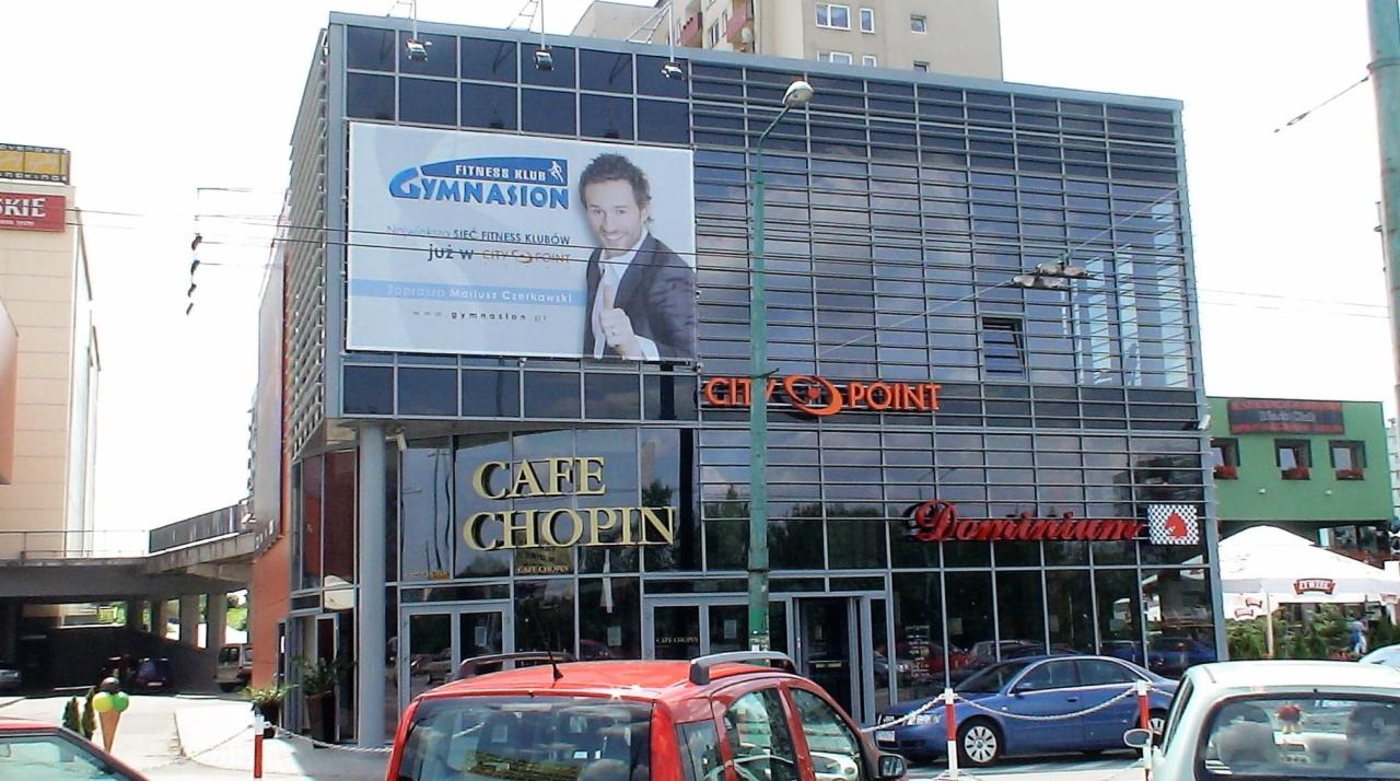 Wielopiętrowy budynek biurowo-usługowy z przeszkloną fasadą, na której widnieją reklamy i szyldy firm, w tym Fitness Klub Gymnasion i Cafe Chopin, z zaparkowanymi samochodami na pierwszym planie.
