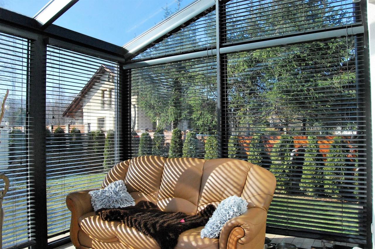 Wnętrze przeszklonego pomieszczenia z aluminiowymi żaluzjami w kolorze antracytu, przez które widać dom i zieleń. Na pierwszym planie skórzana sofa z poduszkami i narzutą, oświetlona promieniami...