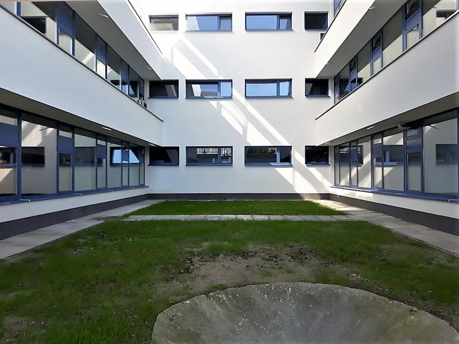 Wewnętrzny dziedziniec budynku z białymi ścianami i regularnie rozmieszczonymi oknami z niebieskimi ramami, trawnik w centralnej części.