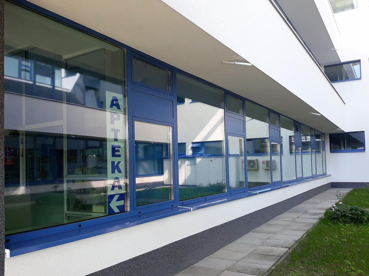 Fasada apteki z niebieskimi oknami aluminiowymi i napisem APTEKA na szybie, widok z chodnika.