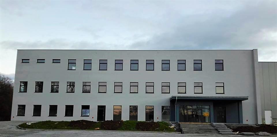 Nowoczesny budynek biurowy z dwoma rzędami okien, świeżo posadzoną zielenią u podstawy i szarym niebem w tle.