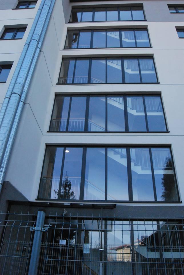 Elewacja budynku z dużymi, prostokątnymi oknami z czarnymi ramami, widoczne schody wewnątrz, zewnętrzna instalacja wentylacyjna i metalowe ogrodzenie.