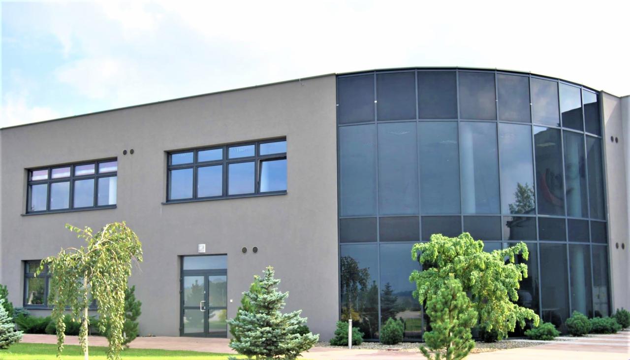 Nowoczesny budynek biurowy z szarymi ścianami i dużymi, ciemnymi oknami, w tym panoramiczne okno narożne, otoczony zielenią.