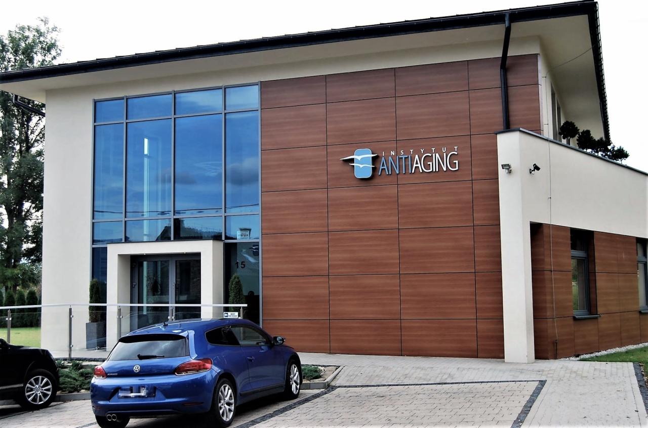 Nowoczesny budynek z dużą szklaną fasadą, brązową elewacją z logo 'Instytut Antiaging' i niebieskim samochodem zaparkowanym przed wejściem.