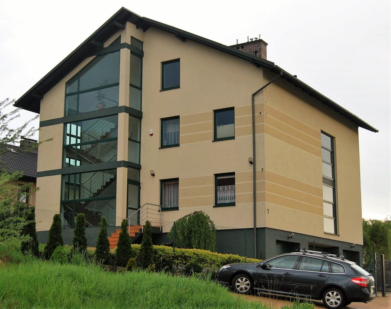 Dwupiętrowy dom z beżową elewacją, dużymi przeszkleniami klatki schodowej z widocznymi schodami, zielonymi obramowaniami okien i ciemnym samochodem osobowym zaparkowanym przed posesją.