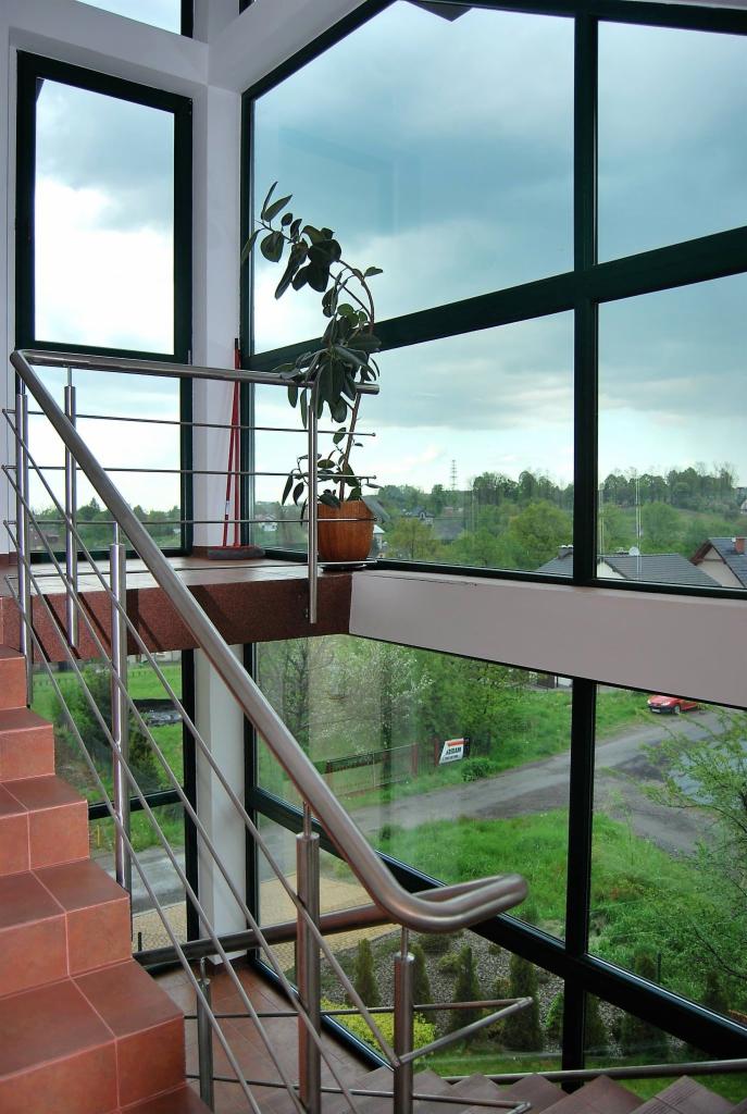 Wnętrze z dużymi oknami PCV z widokiem na zewnątrz, schody z terakoty z metalową balustradą, doniczka z rośliną na podwyższeniu.