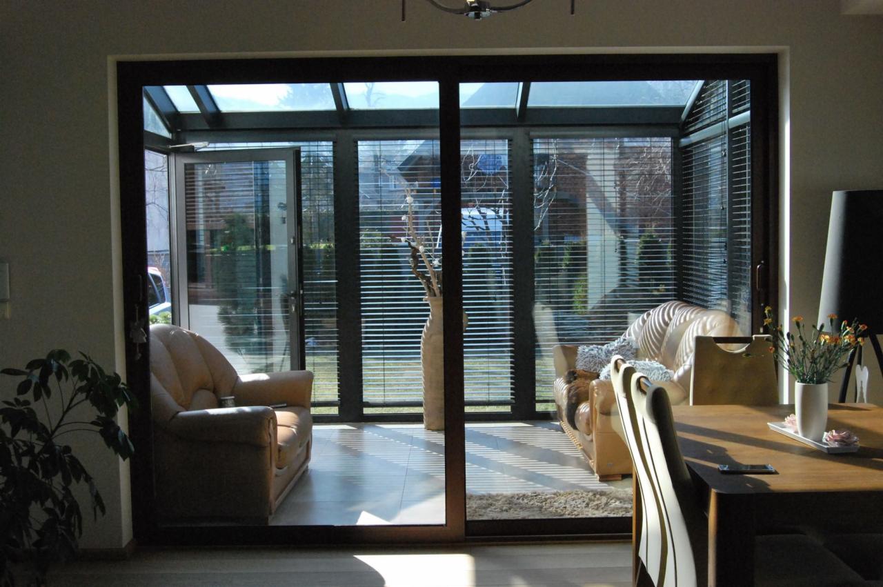 Jasny salon z dużymi przesuwnymi drzwiami tarasowymi i przeszklonym dachem, widok na ogród przez poziome żaluzje.