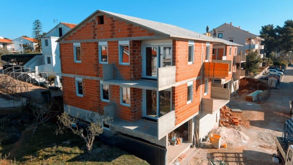 Surowy stan budynku wielorodzinnego z częściowo zamontowanymi oknami i drzwiami balkonowymi, widoczne cegły i beton, robotnicy na dachu.