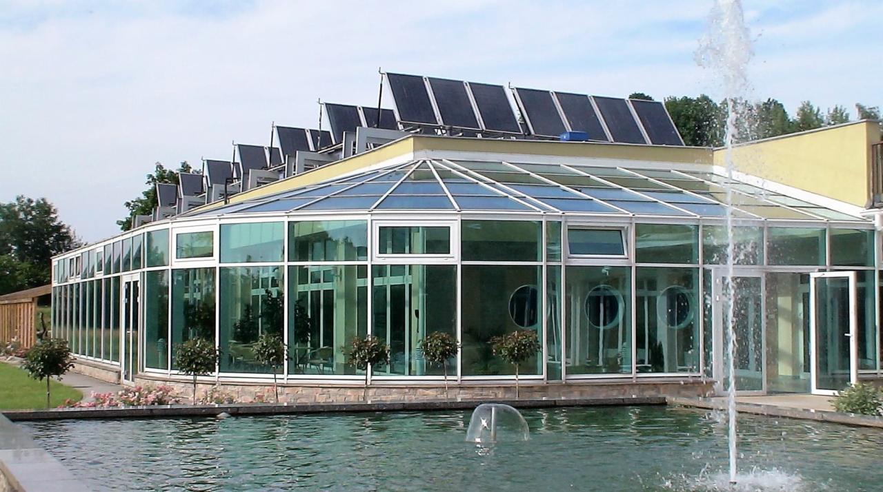 Szklana oranżeria z aluminiowymi oknami, widok z zewnątrz na tle fontanny i paneli słonecznych na dachu.
