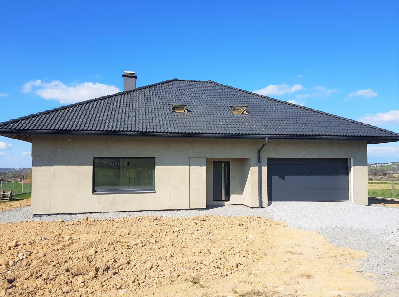 Nowoczesny dom jednorodzinny z grafitową dachówką i ciemnoszarą stolarką okienną, widok z przodu na tle błękitnego nieba. Okno w salonie odbija krajobraz.