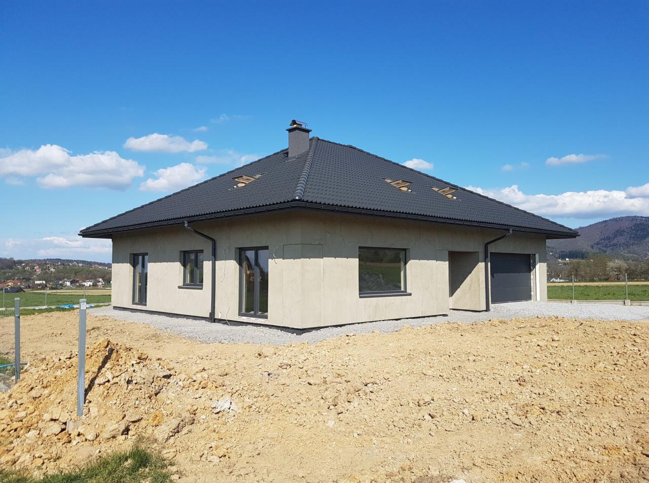 Nowoczesny dom jednorodzinny z szarymi oknami i dachem, widok z zewnątrz na tle błękitnego nieba i wzgórz, teren wokół domu w trakcie zagospodarowania.