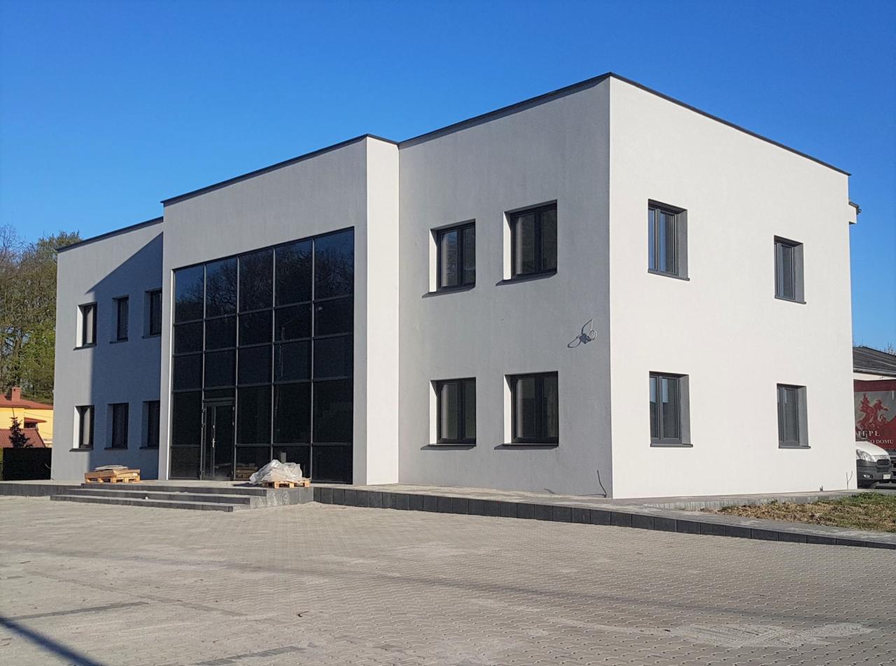 Nowoczesny budynek biurowy z dużą szklaną fasadą, czarnymi ramami okiennymi i minimalistycznym designem, palety z materiałami budowlanymi przed wejściem, błękitne niebo w tle.