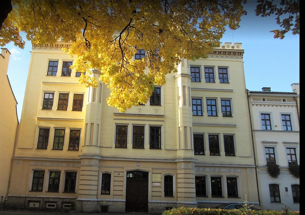 Elewacja odnowionego budynku z żółtą fasadą, widoczna przez gałęzie drzewa z jesiennymi liśćmi, ukazująca rzędy ciemnych okien i ciemne drzwi wejściowe.