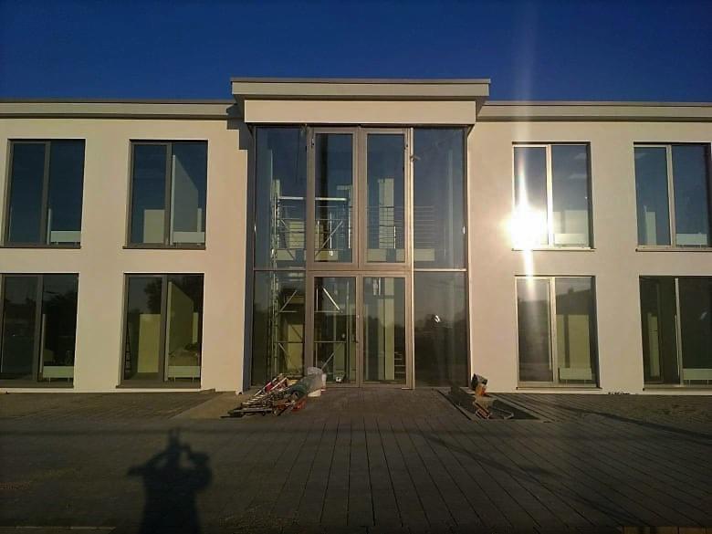 Nowoczesny budynek z dużymi oknami i szklanym wejściem, widoczne rusztowanie wewnątrz, cień osoby w dolnej części kadru.