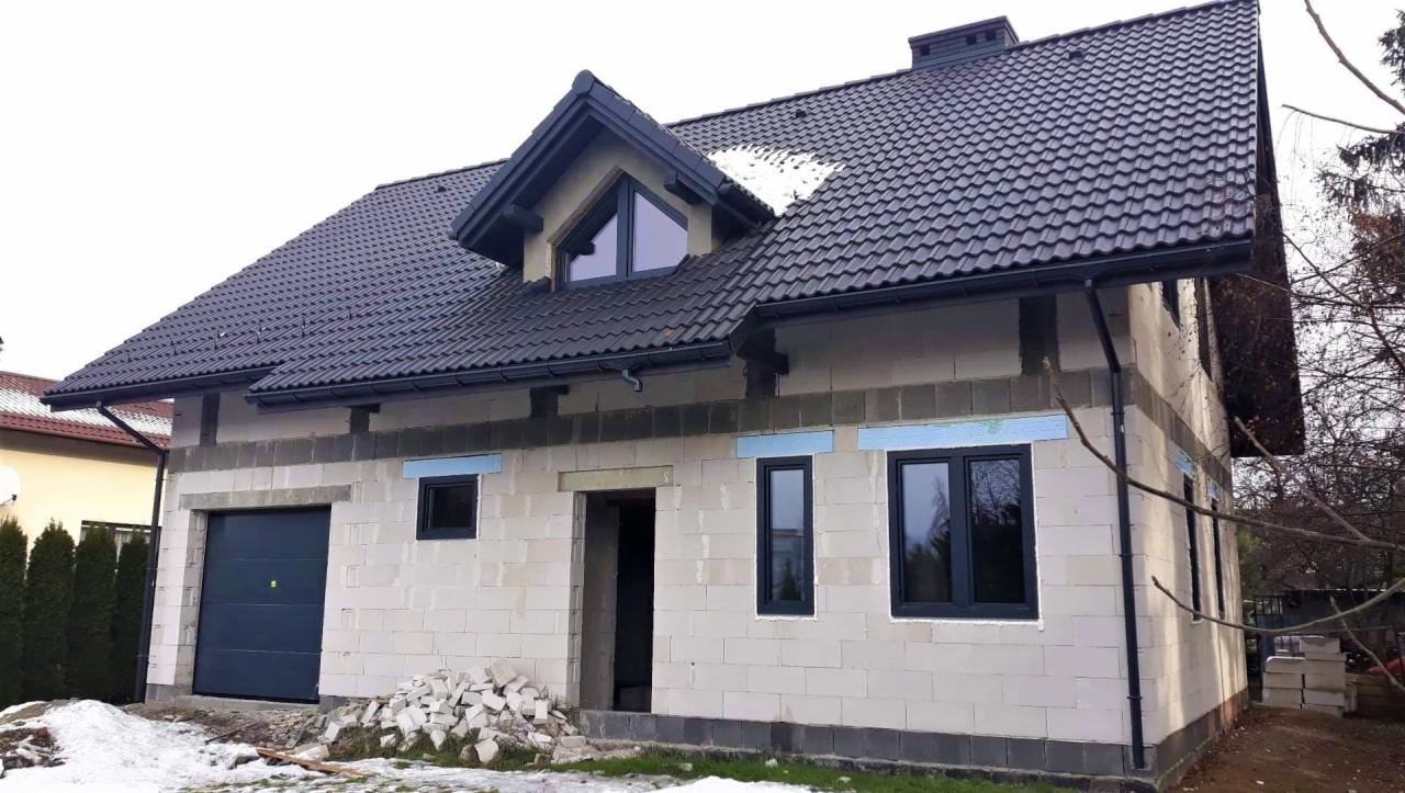 Dom w trakcie budowy z zamontowanymi czarnymi oknami i grafitową bramą garażową, widoczne bloczki betonowe i ocieplenie styropianem na ścianach.