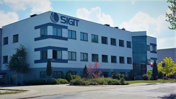 Nowoczesny budynek biurowy z licznymi oknami z ciemnymi ramami, logo firmy na fasadzie, otoczony zielenią i chodnikiem.