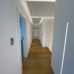 Korytarz z drewnianą podłogą, białymi ścianami i nowoczesnym oświetleniem LED w suficie oraz pionowymi paskami na ścianach, widoczny włącznik światła i białe drzwi w głębi.