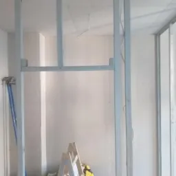 Konstrukcja z metalowych profili, częściowo obłożona płytami, widoczne narzędzia: poziomica, drabina, kable elektryczne wystające ze ściany.