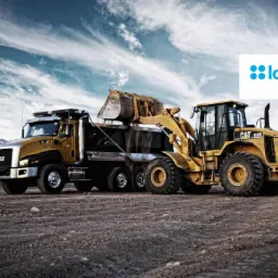 Żółta ładowarka CAT 950H ładuje ziemię do wywrotki na tle gór i pochmurnego nieba, obok logo Idea Getin Leasing.