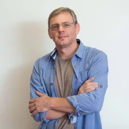 Portret mężczyzny w okularach, z założonymi rękami, ubrany w niebieską koszulę i szary t-shirt, stojącego na tle białej ściany.