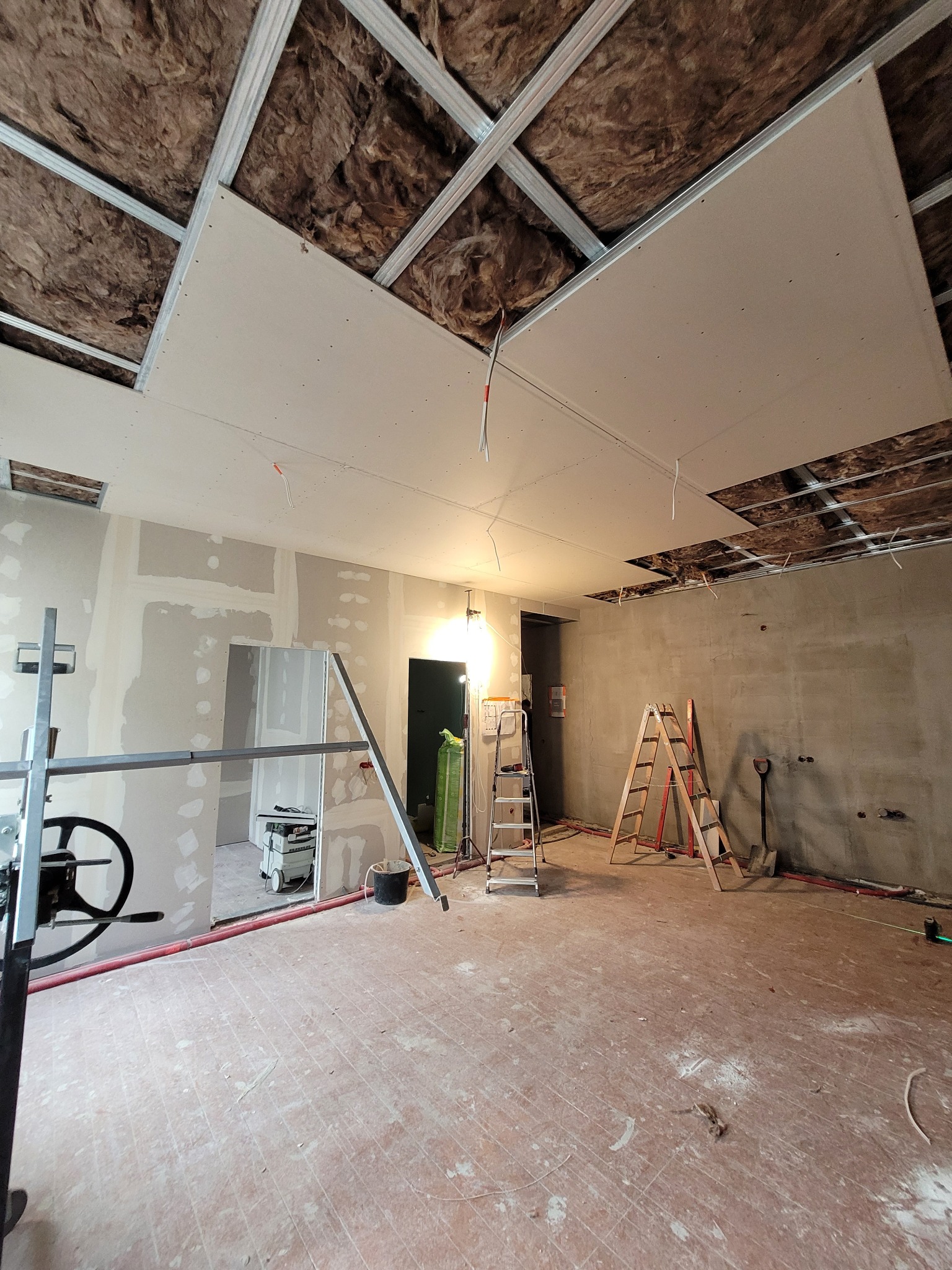 Wnętrze w trakcie remontu: podwieszany sufit z izolacją, ściany w trakcie szpachlowania, drabiny i narzędzia budowlane. Widok na adaptację kamienicy.